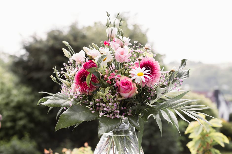 A vibrant floral bouquet arranged in a clear glass vase featuring pink roses, white daisies, and bright pink gerbera daisies accented by lush green foliage, including large leaves and smaller sprigs, 