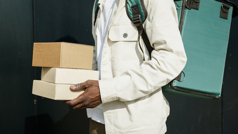 A person wearing a white jacket and carrying a teal backpack is holding three boxes, with the top box being brown and the bottom two boxes being white. The background is dark, contrasting with the per
