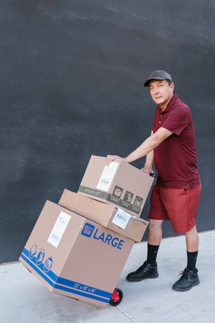 A delivery person is standing beside a dark gray wall, holding a handle on a two-wheeled dolly loaded with two cardboard boxes, one slightly larger than the other. The individual is dressed in a maroo