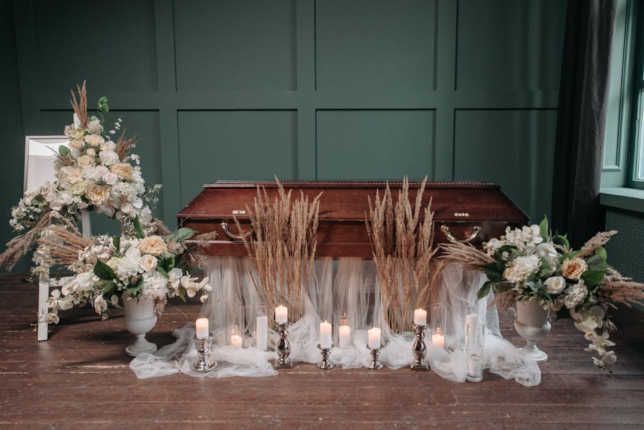 A funeral floral arrangement featuring two large white ceramic vases filled with cream and white roses, hydrangeas, and other soft-colored flowers, accented with pampas grass and green foliage. The va