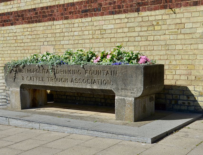 A rectangular stone flower bed attached to a brick wall, filled with a variety of fresh flowers including pink, white, purple, and blue blooms, arranged in a lush, spilling display. The flowers are se