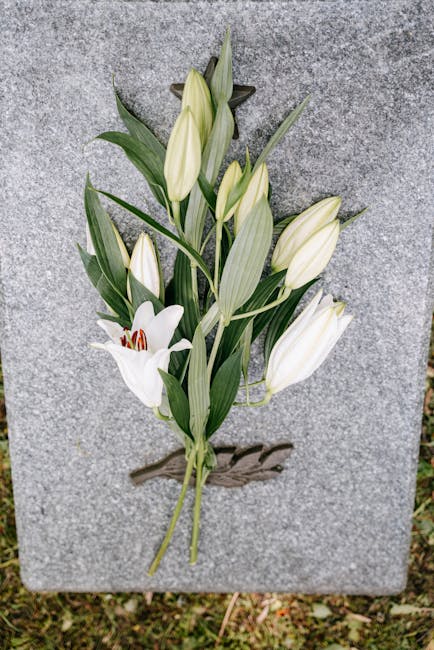 A floral arrangement featuring a bouquet of white lilies and lilies of the valley with green, elongated leaves, laid upon a grey stone memorial surface. The lilies are in various stages of bloom, with